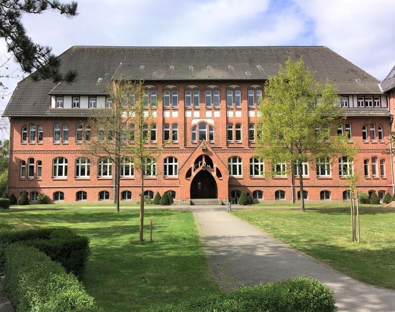 Backsteingebäude mit Spitzdach, symmetrischer Fassade und Bäumen im Vordergrund.