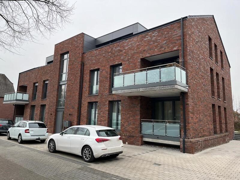 Modernes rotes Backsteinhaus mit Glasbalkonen, zwei weiße Autos, bewölkter Himmel.
