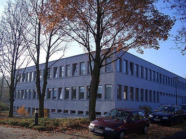 Ein modernes, blaues Bürogebäude umgeben von Bäumen im Herbst mit geparkten Autos vorne.