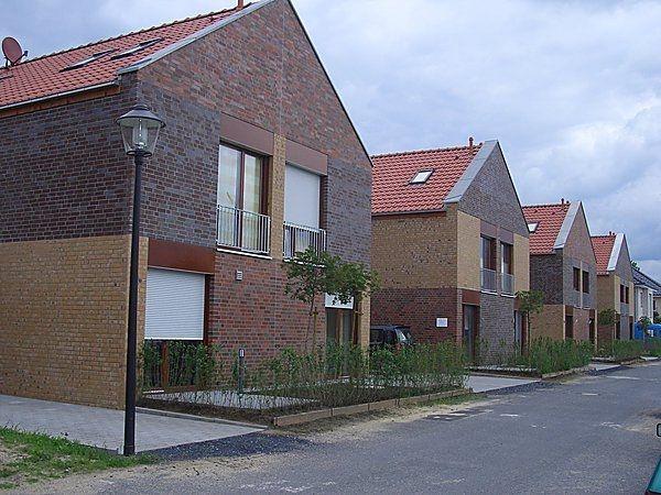 Reihenhäuser aus Backstein, mit roten Dächern und kleinen Gärten; ruhige Straße bei bewölktem Himmel.