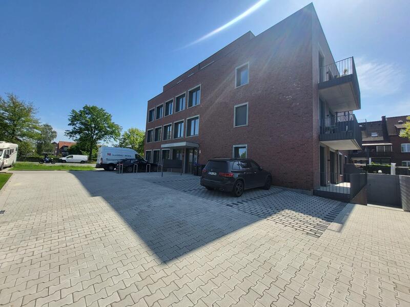 Backstein-Gebäude mit geparktem schwarzen SUV, sonniger Tag, blauer Himmel, grüne Bäume.