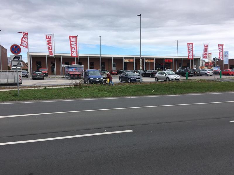 Parkplatz vor einem REWE-Markt mit mehreren geparkten Autos und einer Person mit Kinderwagen.
