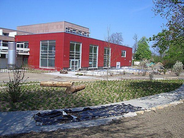 Rotes Gebäude mit großen Fenstern, umgeben von Bäumen und Gehwegen bei klarem Himmel.