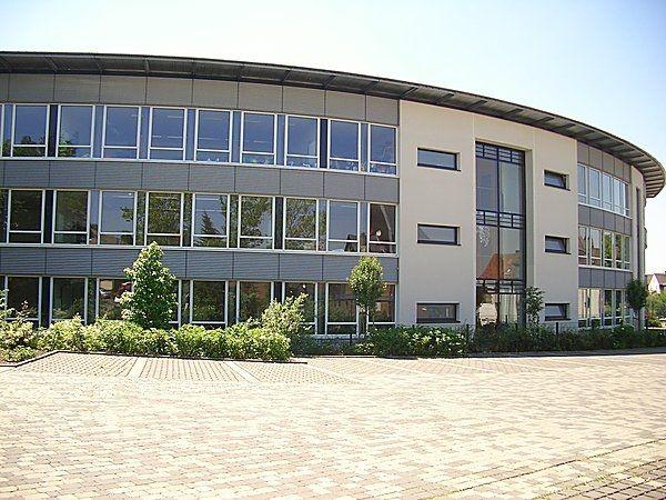 Modernes, geschwungenes Bürogebäude mit grauen Paneelen und großen Fenstern, umgeben von Bäumen.