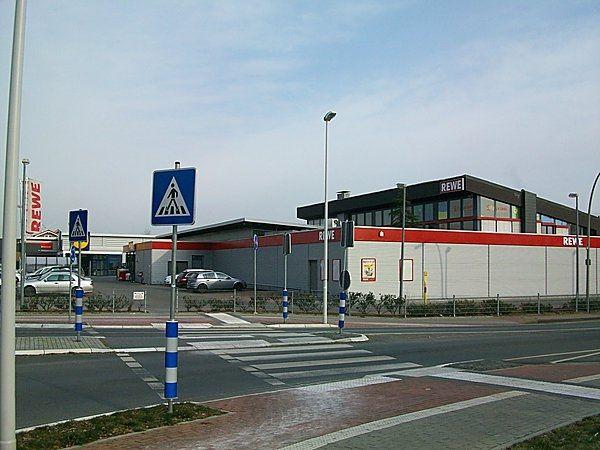 Ein moderner Supermarkt mit roten und grauen Akzenten; Parkplatz und Zebrastreifen im Vordergrund.