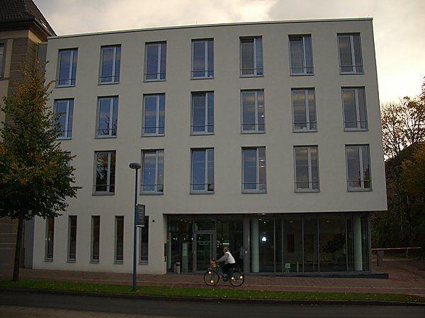 Ein modernes vierstöckiges Gebäude, weiße Fassade, Fensterfront; Person auf Fahrrad fährt vorbei.