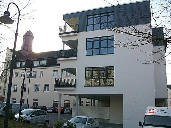 Modernes, mehrstöckiges Gebäude mit Balkonen, weiße Fassade, graue Akzente und Autos im Vordergrund.