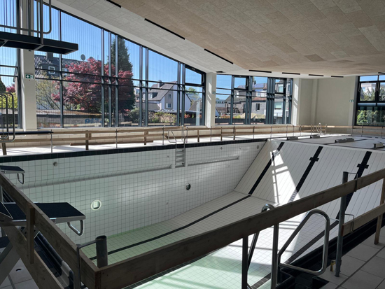 Ein leeres Schwimmbecken mit weißem Fliesenboden und viel Tageslicht durch große Fenster