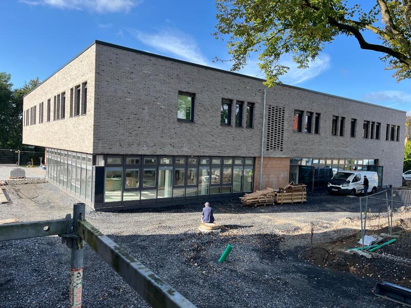 Ein Mann mit dunkler Kleidung sitzt nachdenklich vor einem modernen, hellen Gebäude