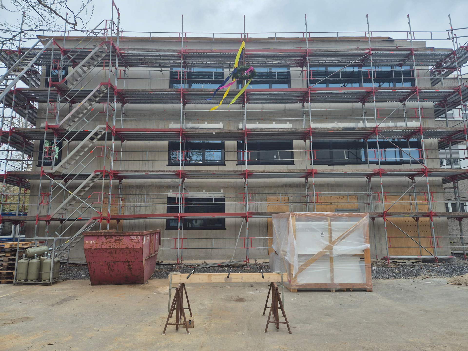 Eine Person mit Leuchtweste und Helm arbeitet lächelnd an einem Gerüst vor Betonfassade