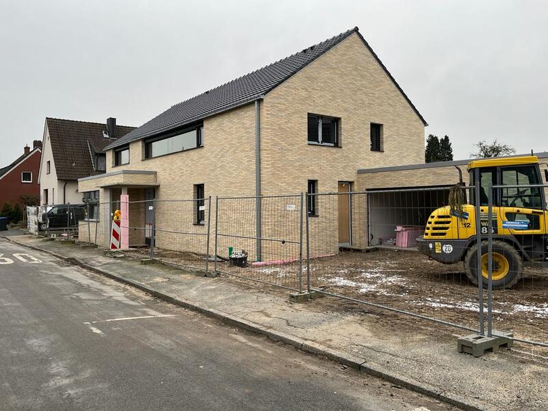 Ein modernes, beigefarbenes Haus im Bau mit Bauzaun und gelber Baumaschine davor