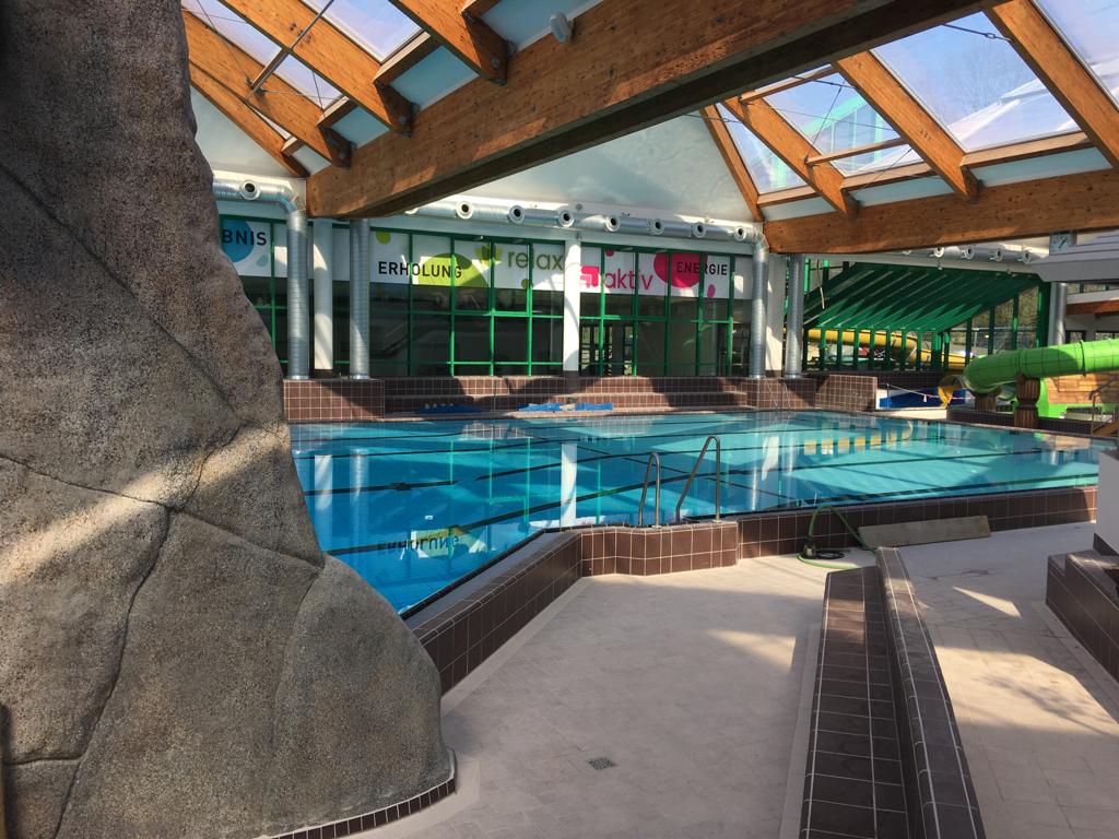 Ein leeres Hallenbad mit Glasdach und Rutsche, Sonnenlicht fällt ins Wasser
