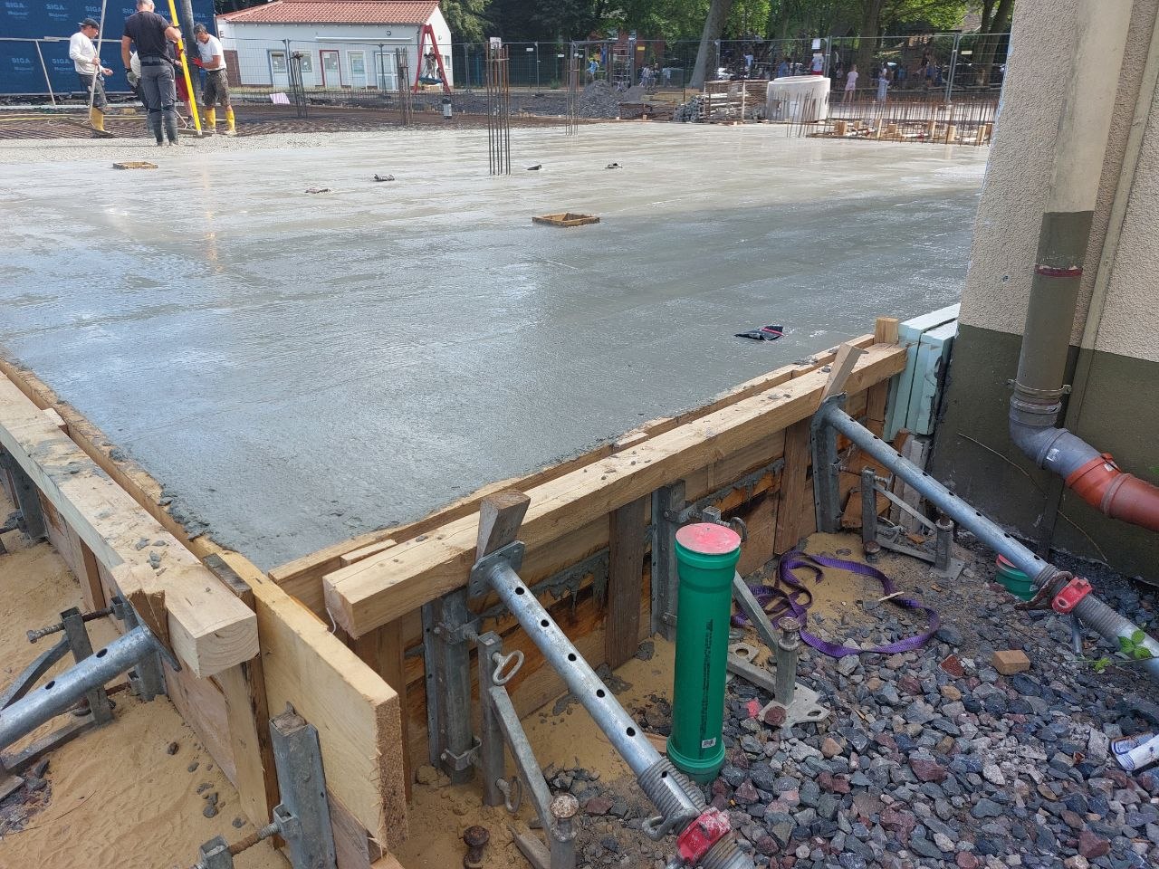 Arbeiter schauen neutral auf graue Betonplatte mit Holzschalung und Rohren